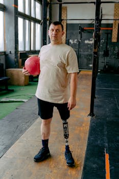 Adult man in gym holding a red medicine ball, showcasing strength and fitness with a prosthetic leg.
