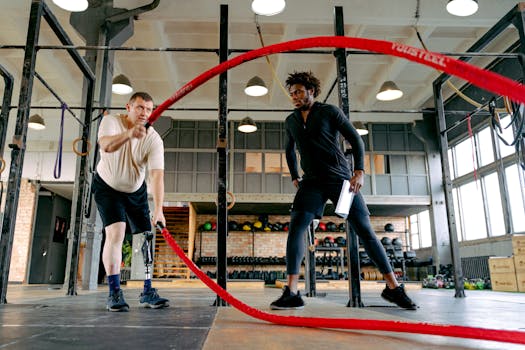 An adult male with a prosthetic leg exercises with ropes, guided by a personal trainer in a gym.