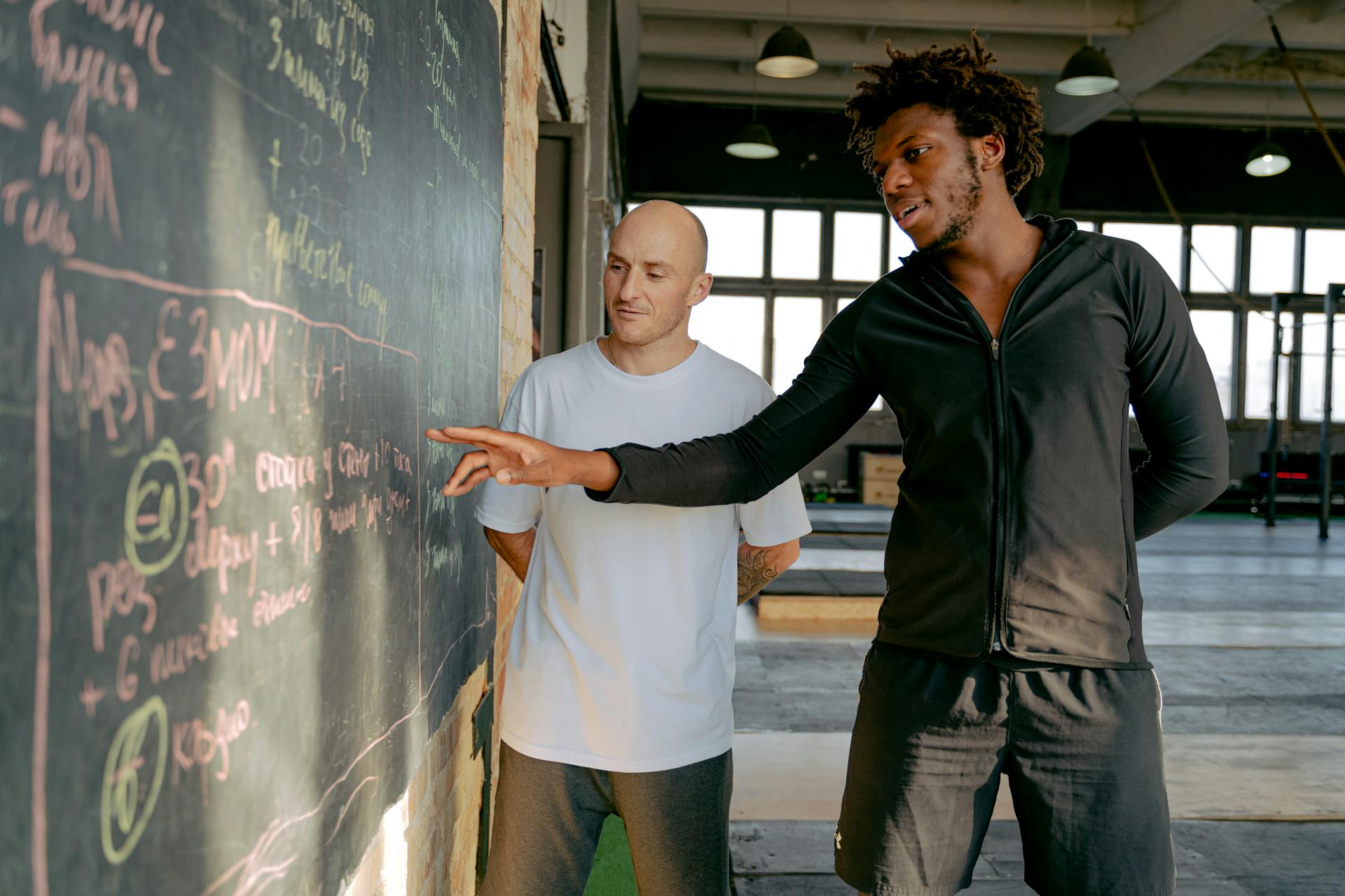 Two men planning a workout strategy on a blackboard in a modern gym setting.