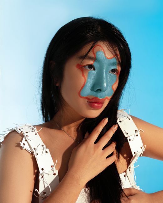 Portrait of Asian woman with painted face looking away putting hand on hair while standing on blue background