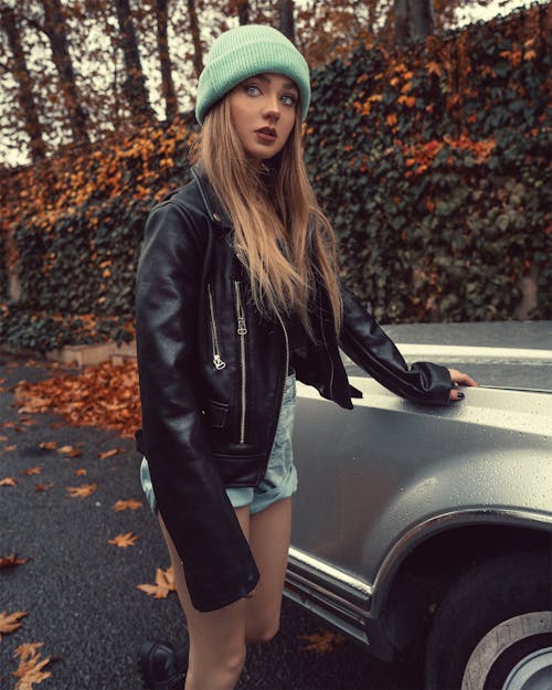 Stylish calm blonde female with long hair in cap and trendy outfit standing near car on road in cold weather in autumn in soft daylight