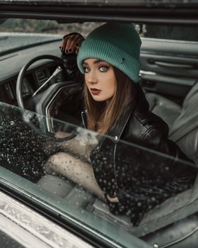 雨天，一位穿着皮夹克、戴着无檐帽的时尚年轻女子坐在车里，流露出自信
