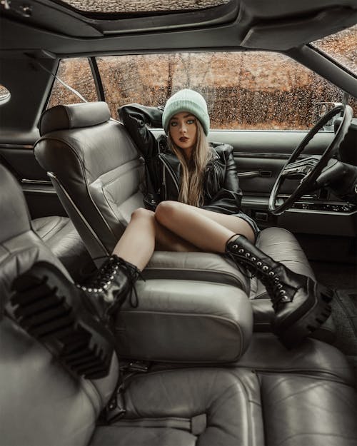 Full body of thoughtful blond woman in cap and stylish black jacket and boots sitting in car on drivers seat near wheel in autumn in daylight