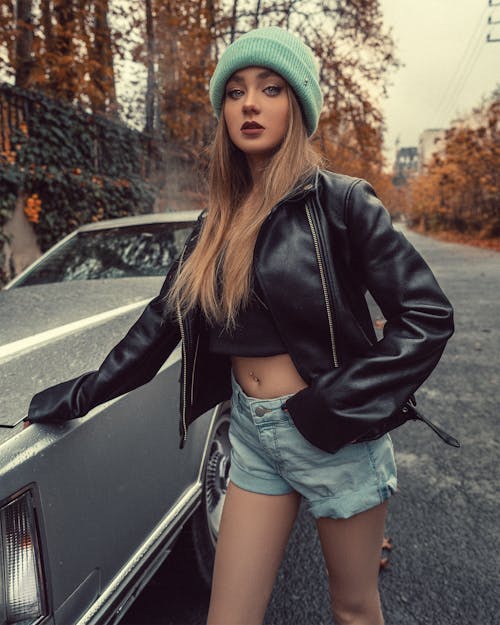 Stylish young female in black leather jacket and shorts standing on asphalt roadway near gray car in cold weather in autumn in soft daylight