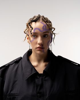 一位扎着辫子、脸上涂着紫色颜料的年轻女模特，穿着宽松的黑色夹克，冷静地站在白墙前，目光移向别处