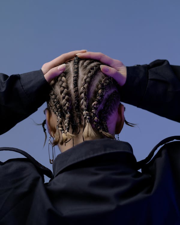 Free Back view unrecognizable female with African braids hairdo wearing black jacket touching head while standing against dark blue wall in studio Stock Photo