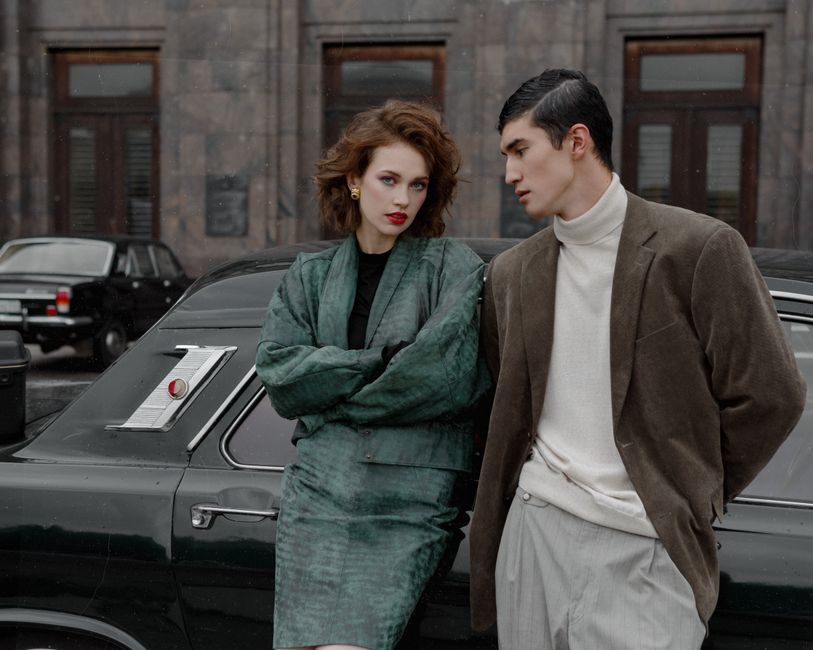 Young stylish woman in elegant green suit with ethnic man in brown jacket standing near black car and looking at camera in soft daylight