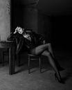 Black and white of young melancholic female model in trendy apparel leaning with hand on table while sitting with crossed legs and looking away