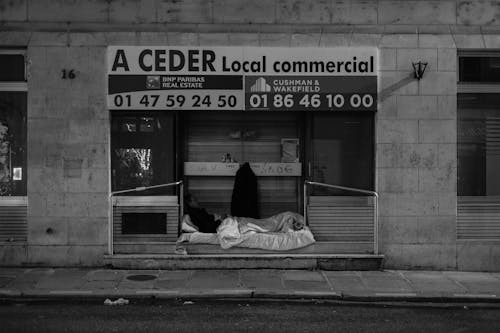 Black and white of homeless man lying on mattress with blanket placed on street near building in city in daytime