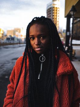 A fashionable woman with braided hair wearing a red coat in a cityscape.
