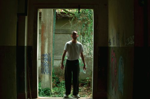 Free A man standing in a doorway of an abandoned, graffiti-covered building with overgrown plants. Stock Photo