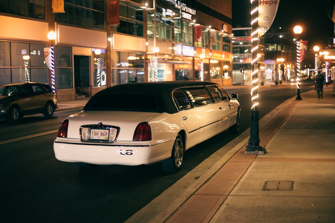 White limo with tinted glasses and license plate on roadway against buildings illuminated by street lights in evening city