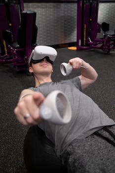 Person using virtual reality headset and controllers for a workout in a gym setting, lying on an exercise ball.