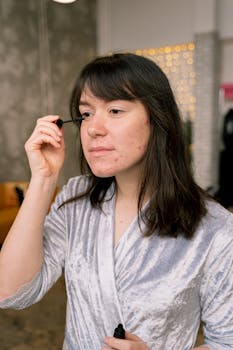 Teen girl applying mascara indoors, wearing a casual robe, focusing on her reflection.