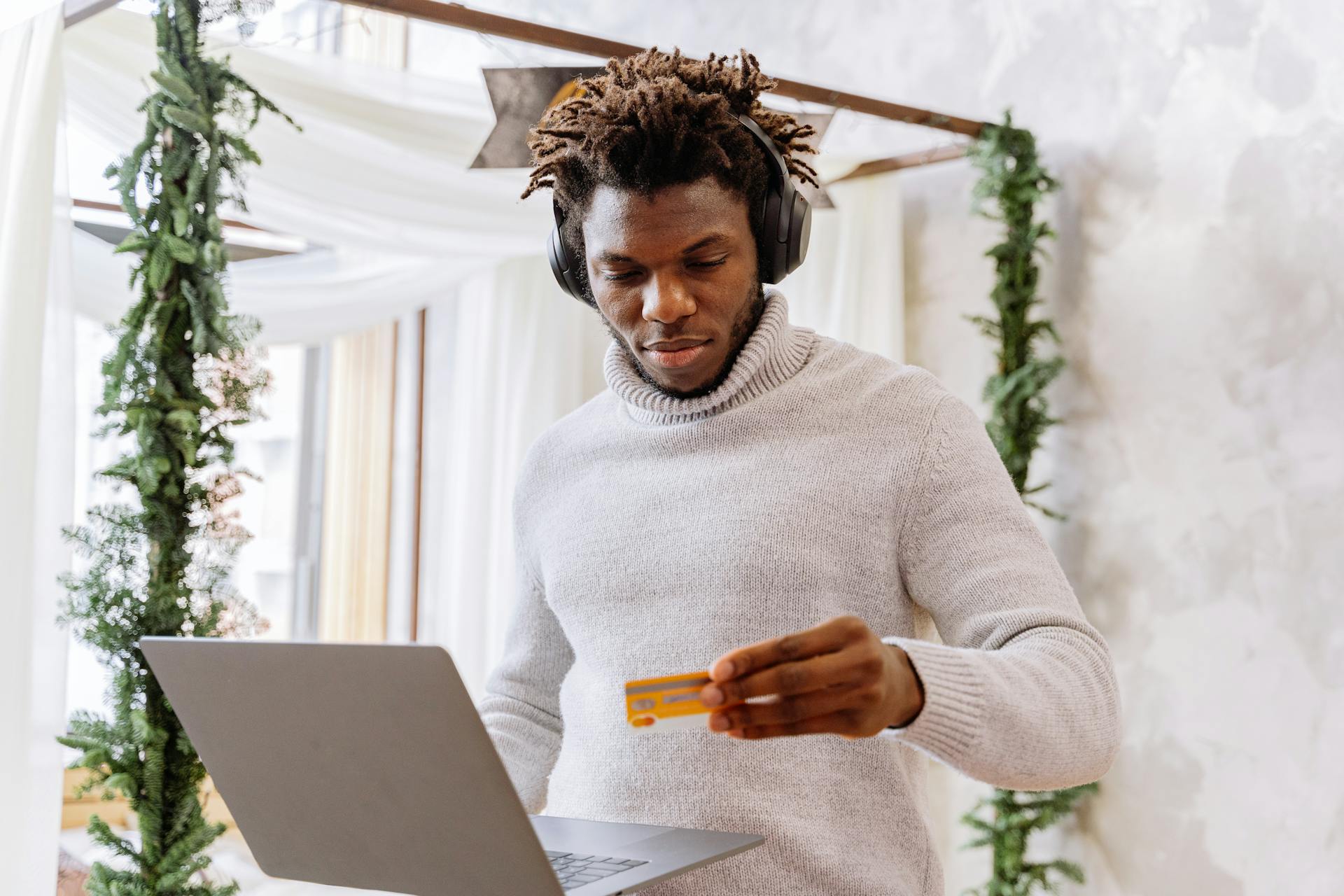 A man stands indoors with a credit card and laptop, wearing headphones and a turtleneck sweater.