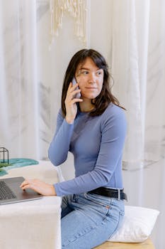 Casual young woman in blue long sleeve and jeans using phone and laptop indoors.