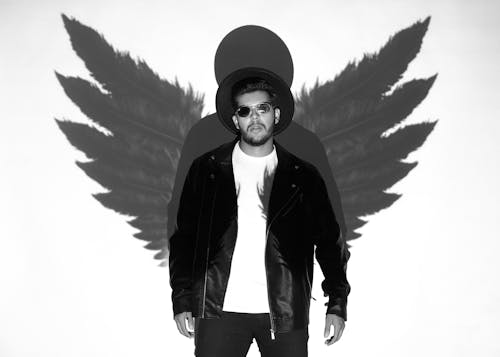 Stylish guy in fashionable sunglasses and hat standing near white wall with angel wings shadow