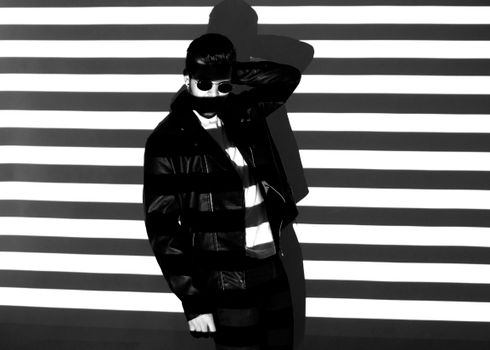 Black and white of unrecognizable bearded guy in trendy leather jacket and sunglasses standing near wall with jealous shadow in sunlight