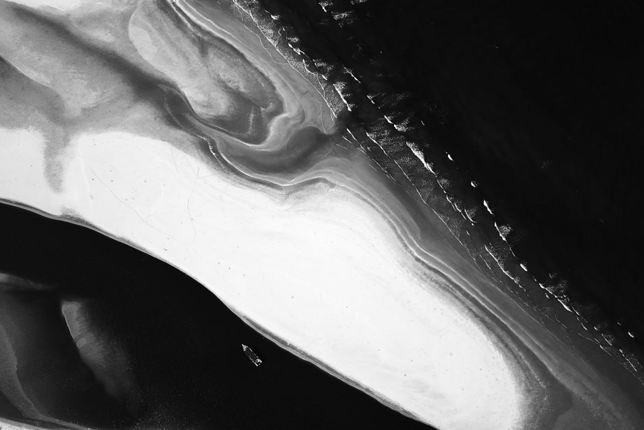 Black and white aerial view of island with wavy surface between ocean with sailing boat
