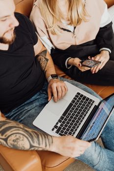 A man with tattoos and a woman use a laptop and smartphone on a couch indoors.