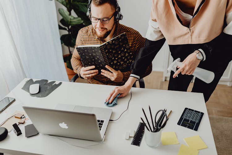 Woman Cleaning The Desk While Man Sits And Uses Laptop 