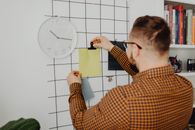 Person arranging notes on an office wall grid beside a clock, creating an organized workspace.