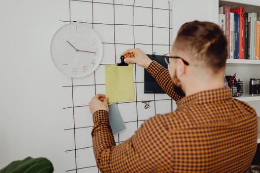 Person arranging notes on an office wall grid beside a clock, creating an organized workspace.