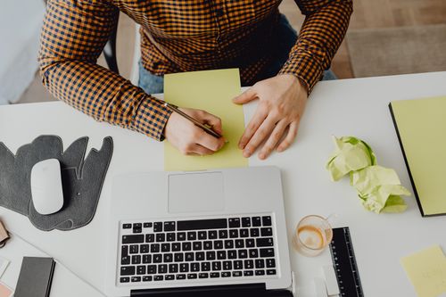 Free A man in a checkered shirt writing on paper at a desk with a laptop and coffee. Stock Photo