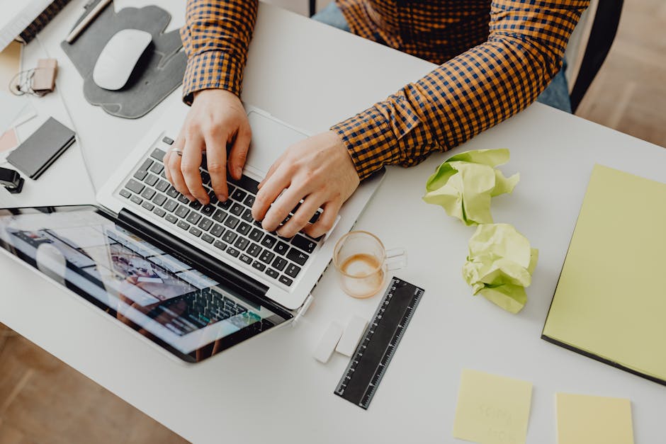 Overhead view of a person typing on a laptop in an office setting with crumpled papers and stationery.
