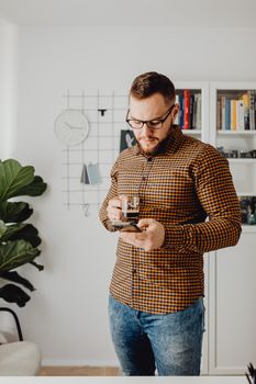 A man in a checkered shirt uses a smartphone while holding a coffee indoors.