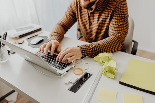 A man wearing a checkered shirt types on a laptop at his office desk.