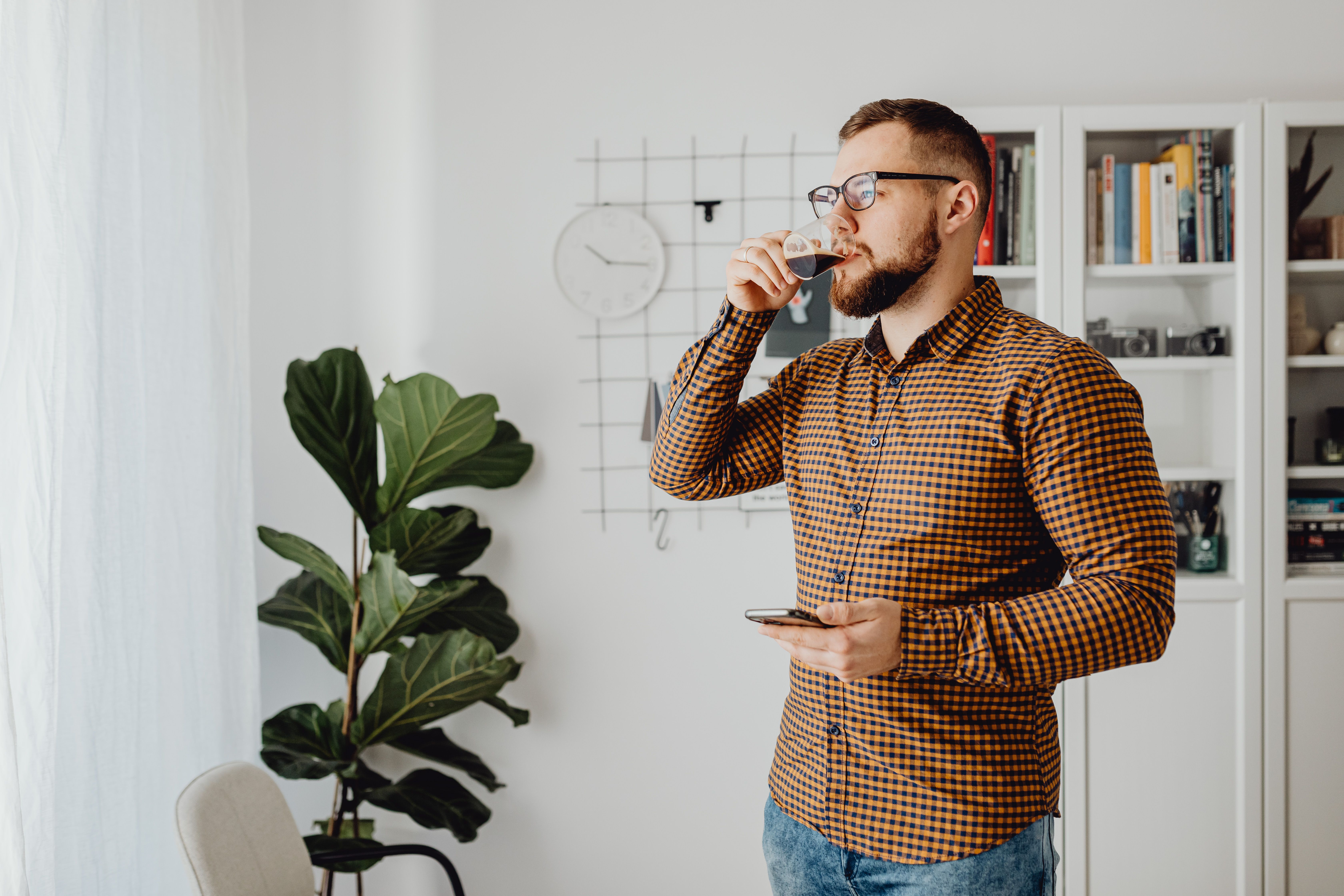 A bearded man with eyeglasses drinks coffee indoors while checking his smartphone.