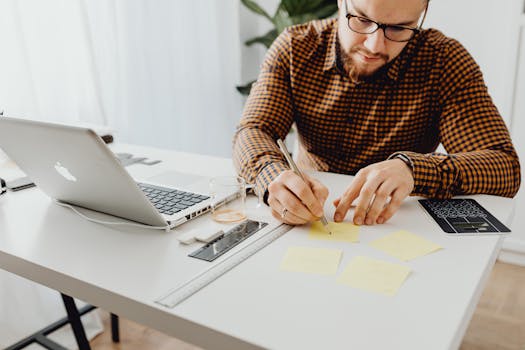 A man with glasses writes notes at his office desk with a laptop and coffee.