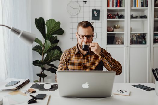 Man with a beard and eyeglasses working on a laptop, drinking coffee in a home office.