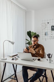 A man with glasses and a beard uses a smartphone at his desk in a bright, modern office setting.