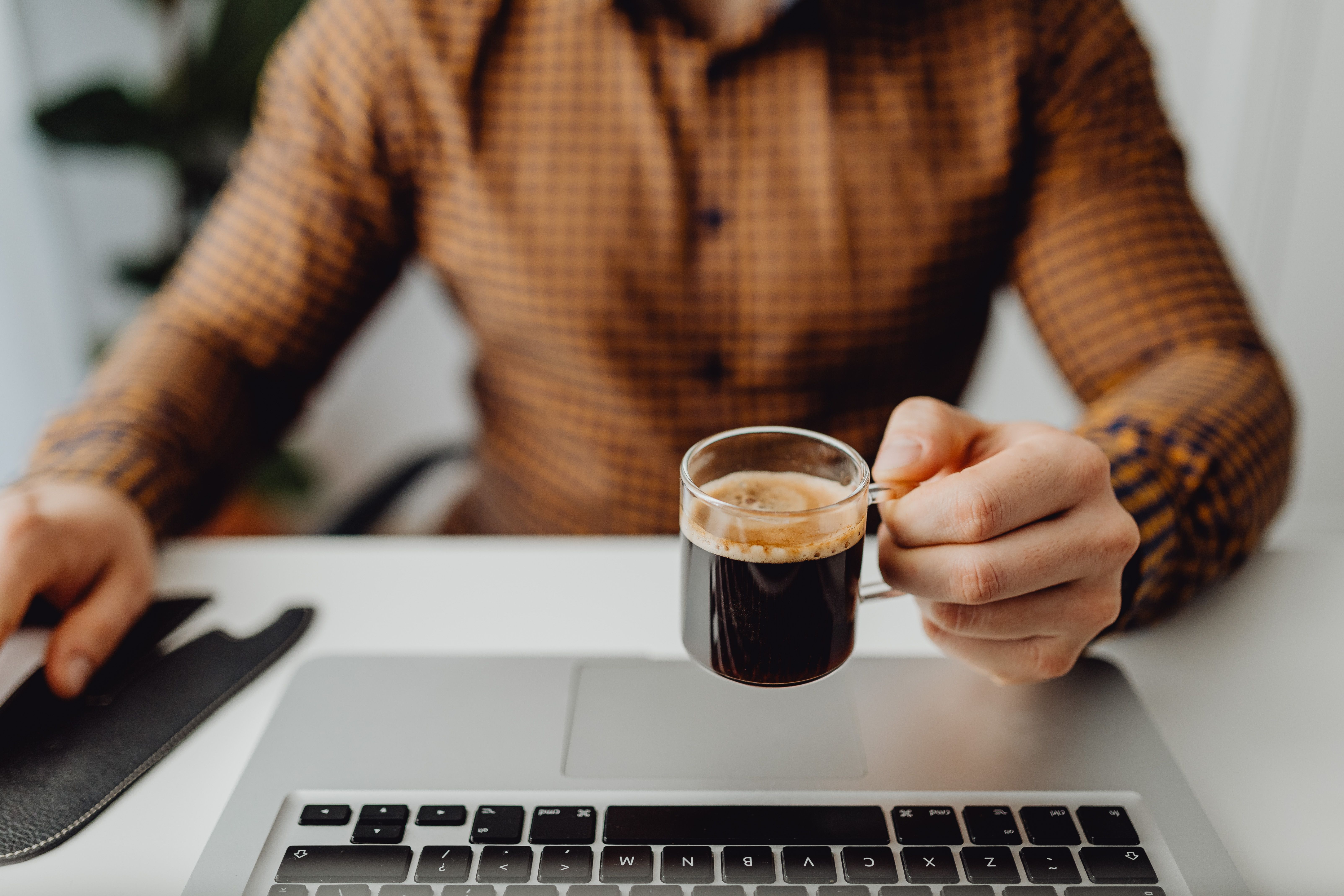 Adult man in checkered shirt holding espresso cup while working on laptop in office setting — modafinil vs caffeine