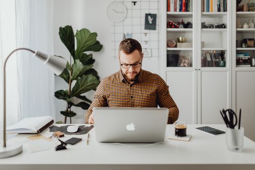 Free Man working on laptop in a modern home office space with plants and bookshelves. Stock Photo