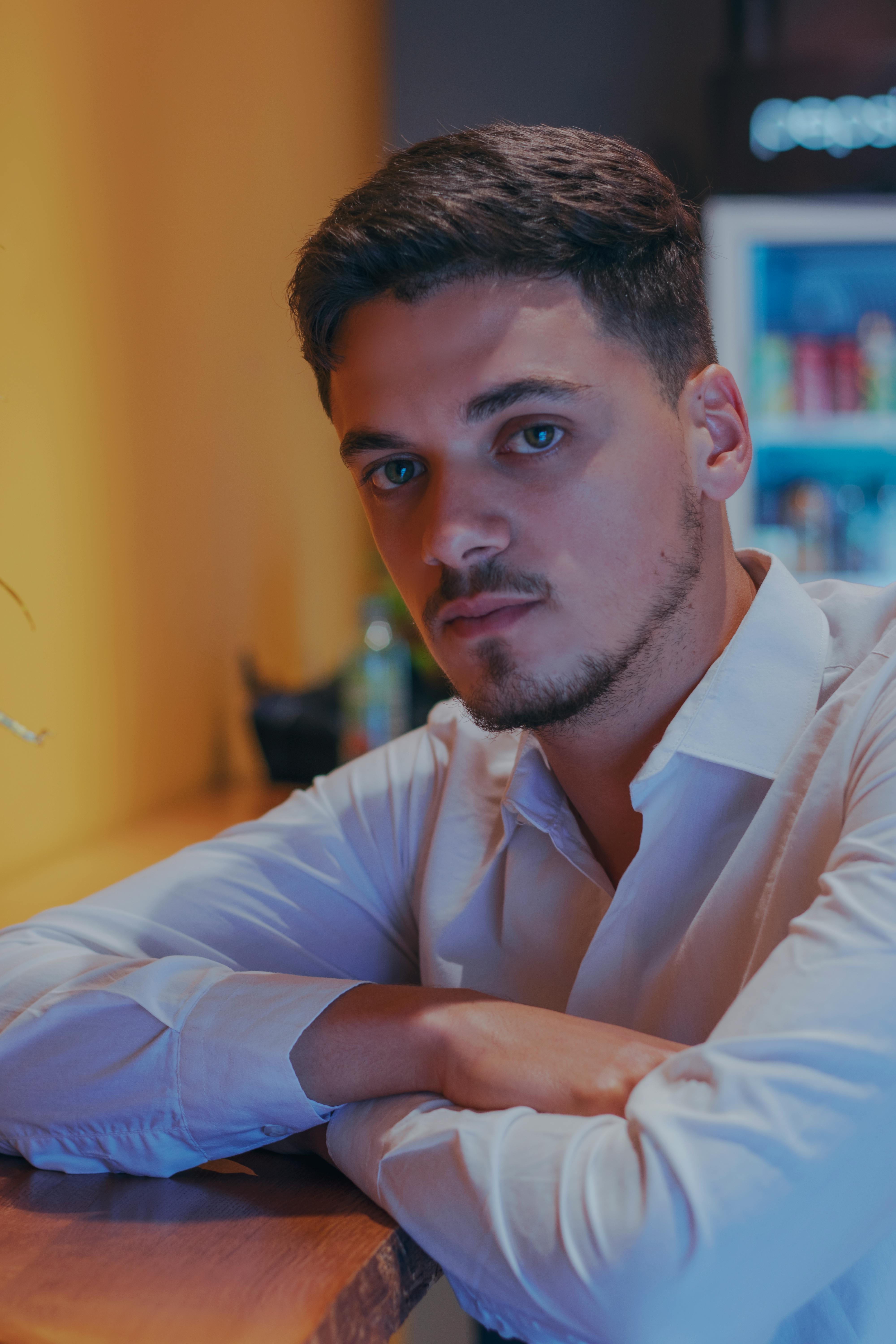 Portrait of a young man with a beard sitting indoors wearing a white shirt.