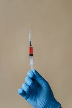 Close-up of a gloved hand holding a syringe with red liquid, against a neutral background.