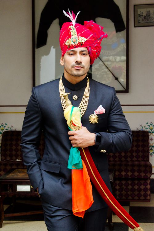 Free Portrait of a stylish Indian man in a black suit and vibrant Rajasthani turban standing indoors. Stock Photo