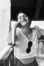 Black and white of content young female in casual clothes smiling and leaning on hand while standing with eyes closed in summerhouse or terrace