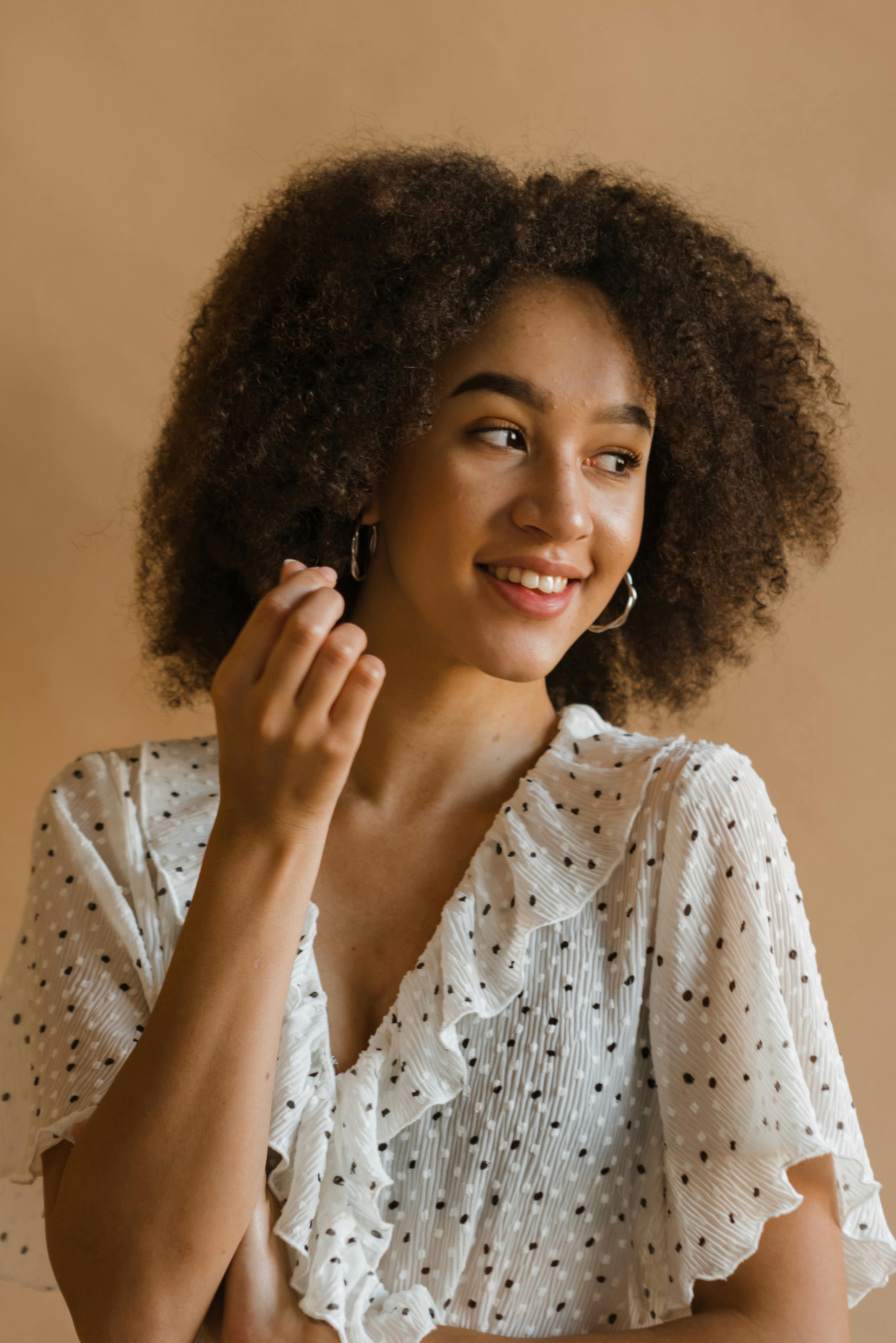 一位快乐的年轻非裔美国女性，卷发，身着浪漫连衣裙，站在米色背景前微笑并移开目光