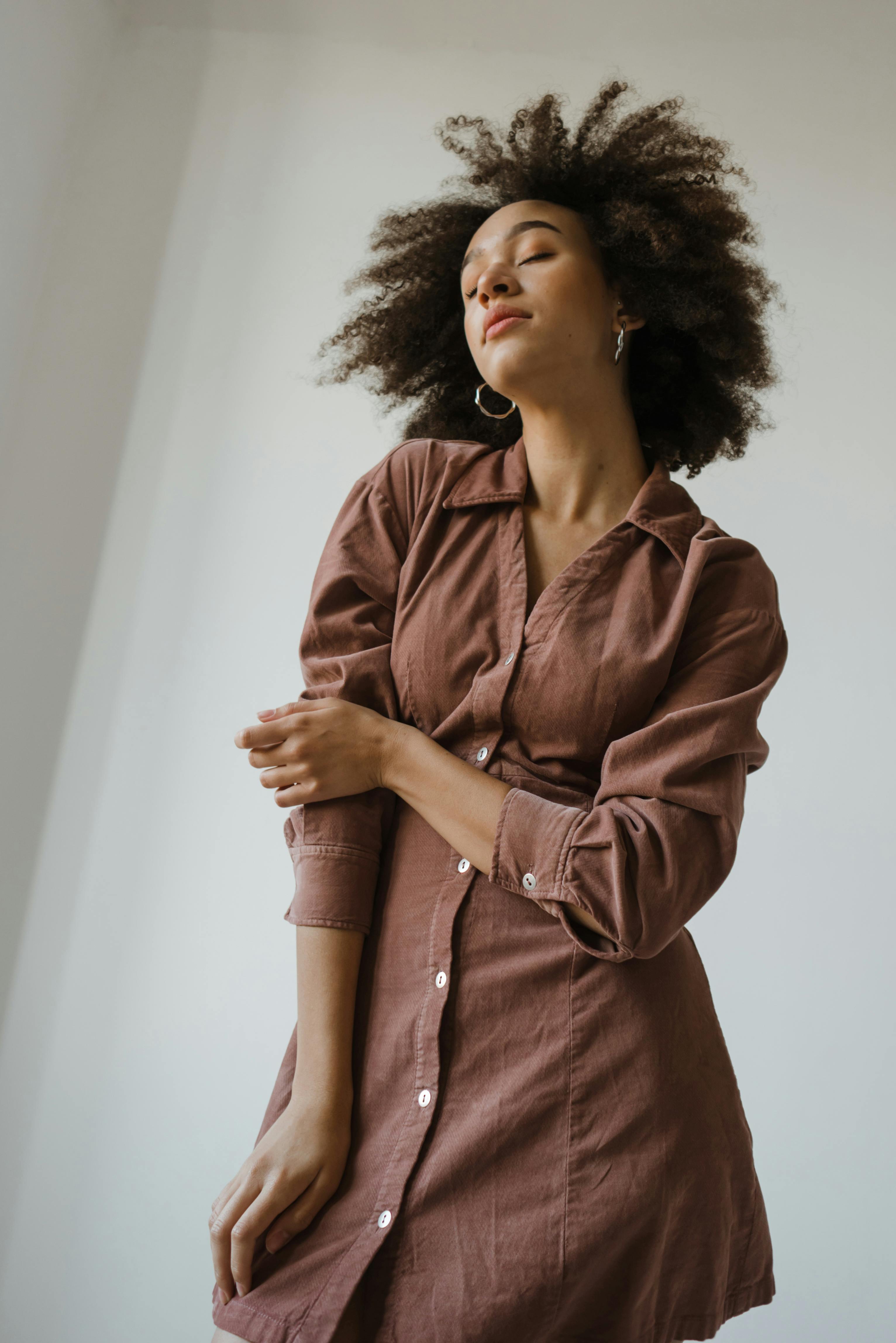 Fashionable young black woman with Afro hair in casual dress standing against white background with closed eyes