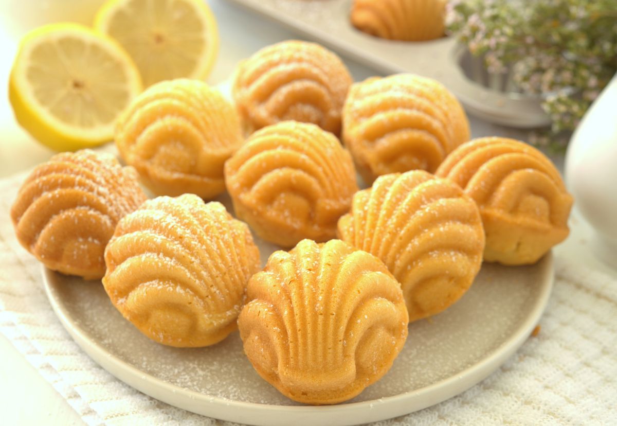 Plate of golden shell-shaped madeleines lightly sugared, with lemon slices and a madeleine pan