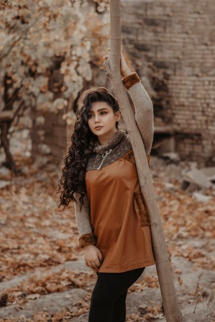 Gentle content female in trendy outfit leaning on trunk of tree on blurred background of yard with autumn leaves