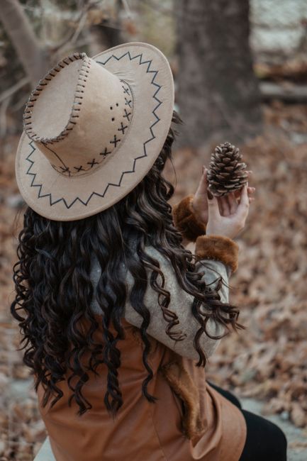 Back view of anonymous female with long wavy hair touching cone in hands on blurred background of autumn leaves