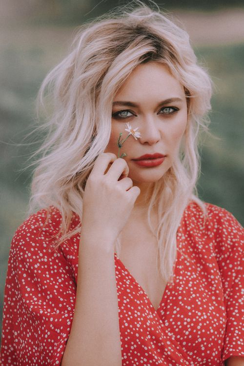 Free Stylish blond female wearing red polka dress looking at camera while standing with small flower in countryside on blurred background Stock Photo