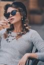 Confident female in sunglasses and black beret sitting on chair and smoking cigarette on street in city on blurred background