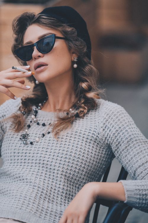 Free Confident female in sunglasses and black beret sitting on chair and smoking cigarette on street in city on blurred background Stock Photo
