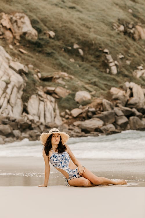 Young feminine woman in modern swimsuit resting on sandy ocean coast against rough mount and looking at camera